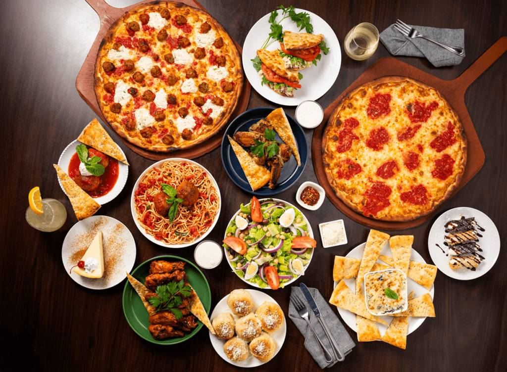 An assortment of Italian dishes, including two pizzas, pasta, salad, wings, and desserts, on a dark wooden table.