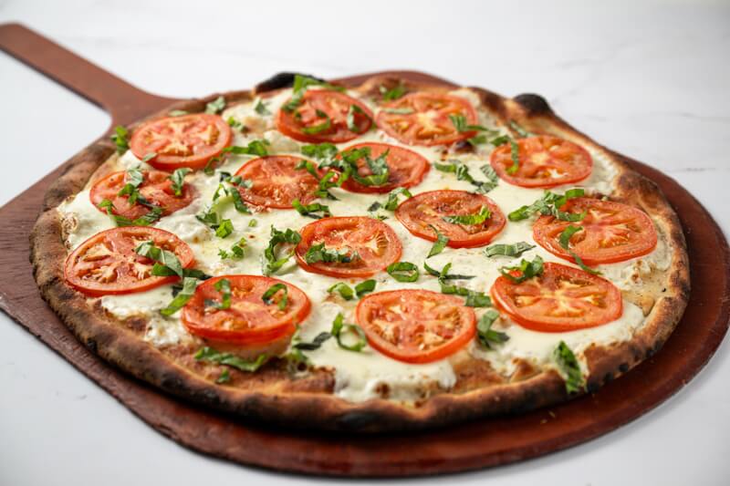 Caprese style pizza featuring sliced tomato, basil, and fresh mozzarella on a coal fired crust with a light, rustic presentation.