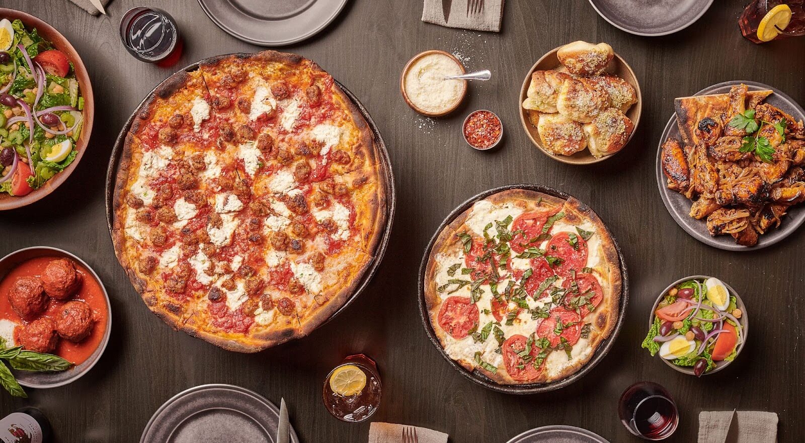 Two pizzas with ricotta, tomatoes, and basil, surrounded by salad, meatballs, garlic knots, wings, and drinks on a dark table.