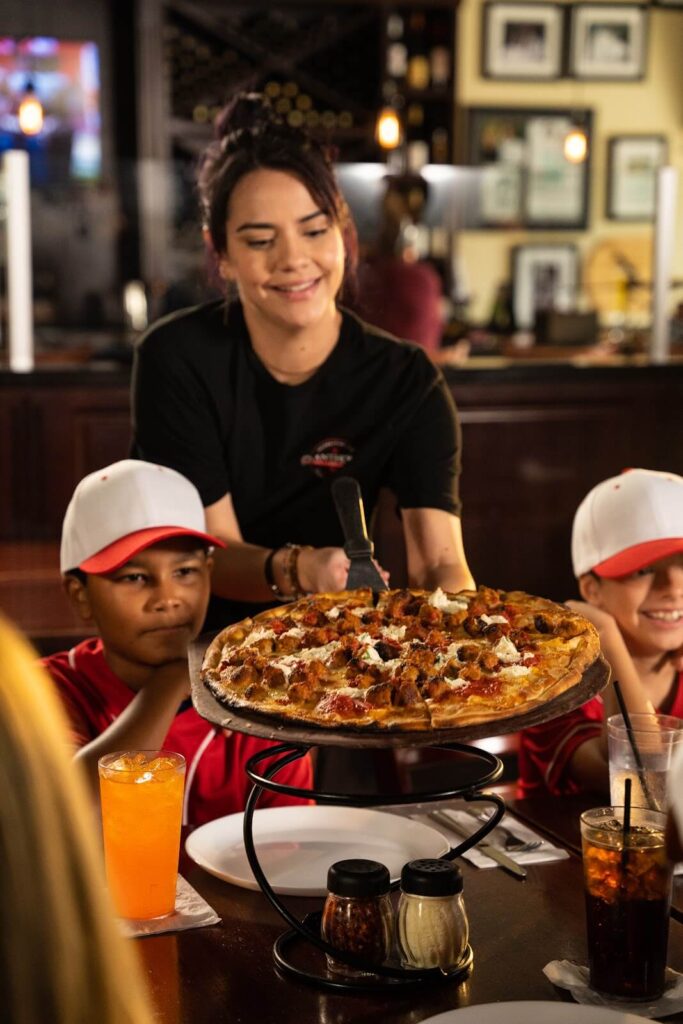 Waitress serving a freshly baked meatball pizza to two young baseball players at a restaurant.