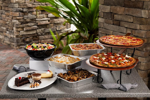 A variety of Italian dishes including pizza, lasagna, wings, pasta, salad, and desserts displayed on a stone table outdoors.