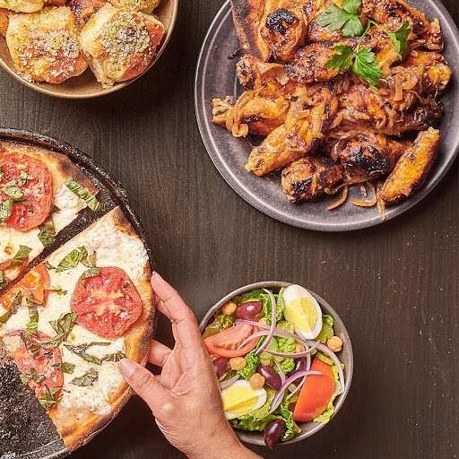 Hand reaching for a slice of Margherita pizza, surrounded by buffalo wings, garlic knots, and a fresh Greek salad on a wooden table at Anthony's.