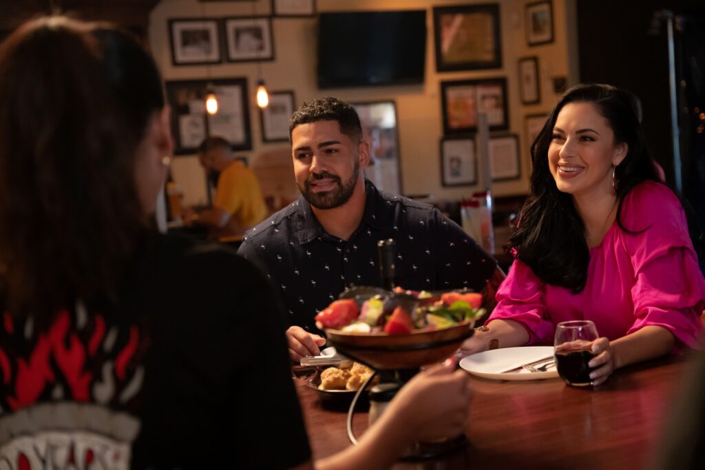 A couple at a restaurant table smiling while talking to a server, with a fondue dish in front.