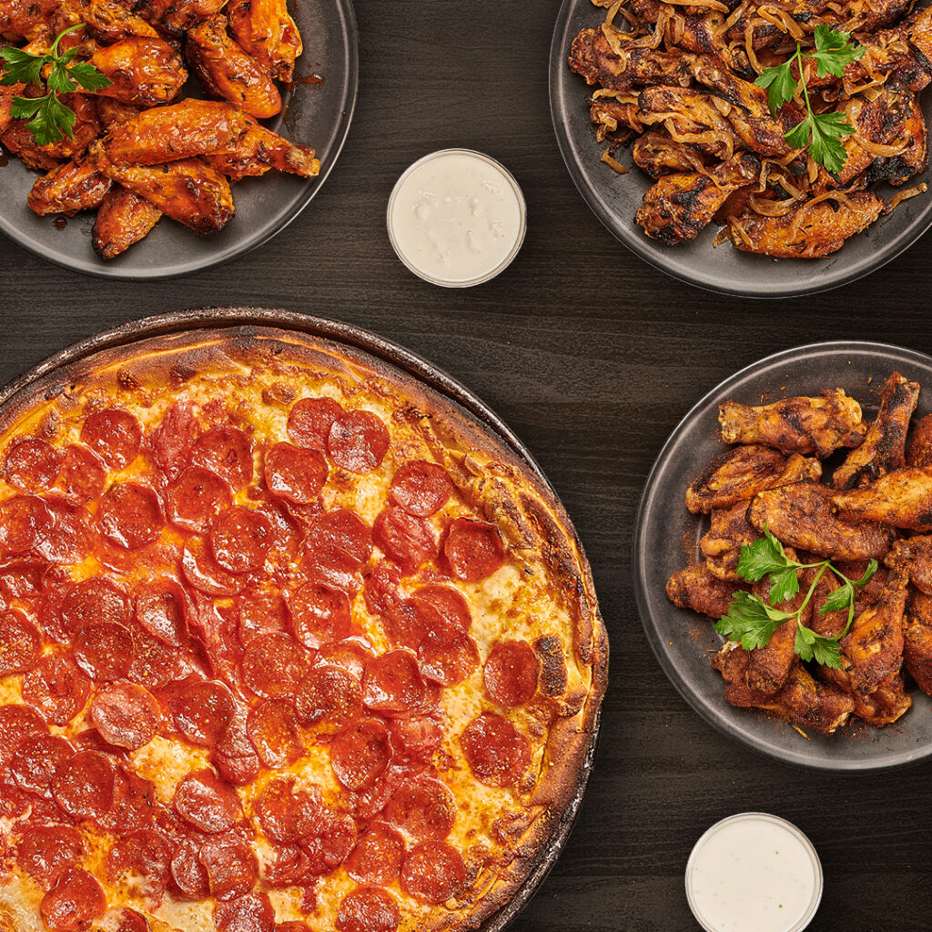 Pepperoni pizza with assorted chicken wings and dipping sauce on a dark table.