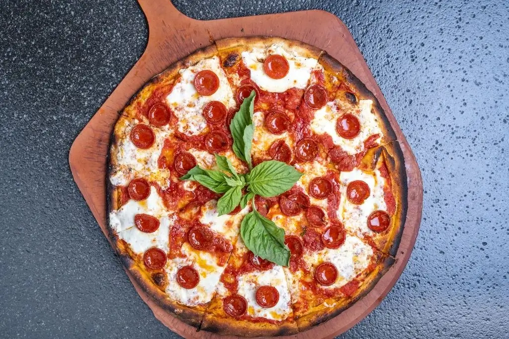 Wood-fired pepperoni pizza with melted mozzarella, fresh basil, and crispy crust on a wooden pizza peel.