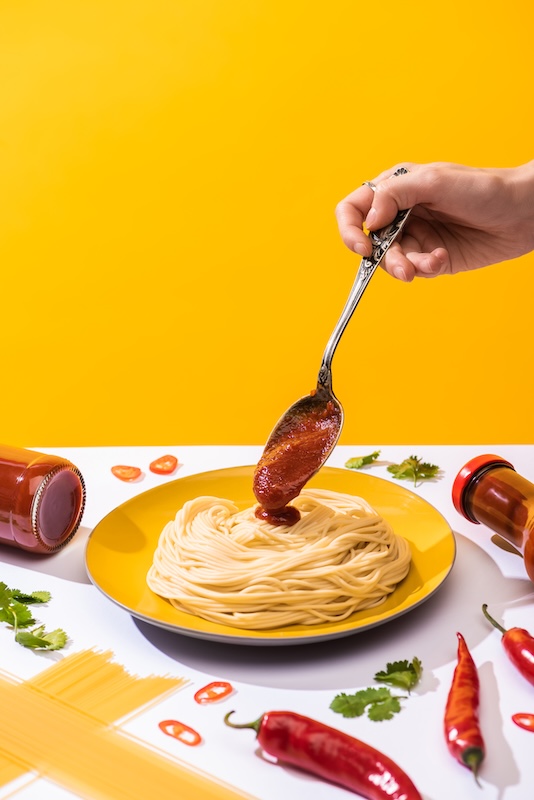 Spaghetti on a yellow plate with tomato sauce being poured from a spoon, surrounded by chili peppers and fresh herbs.