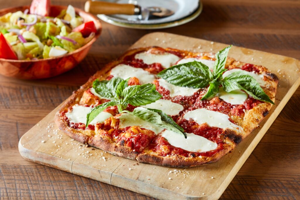 Freshly baked Margherita pizza with melted mozzarella, tomato sauce, and basil on a wooden cutting board, served with a side salad.