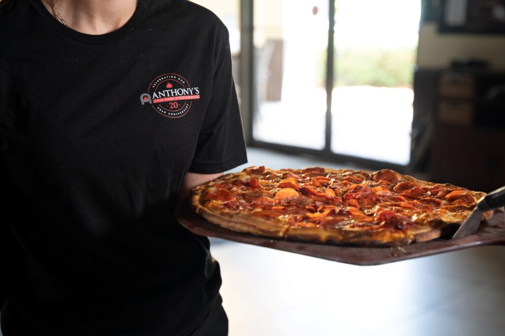 Server in an Anthony’s Coal Fired Pizza shirt holding a crispy, coal-fired pepperoni pizza on a wooden paddle.