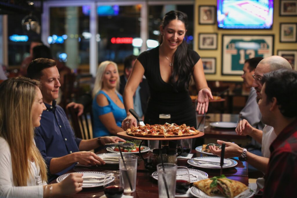 Smiling waitress serving a fresh pizza to a group of friends at a lively restaurant with sports memorabilia on the walls.