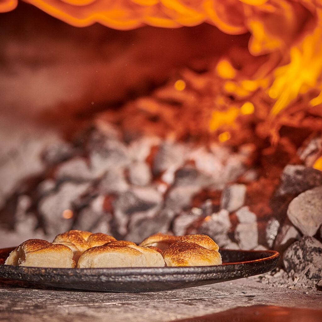 Wood-fired bread rolls on a black plate near hot embers in a traditional oven.