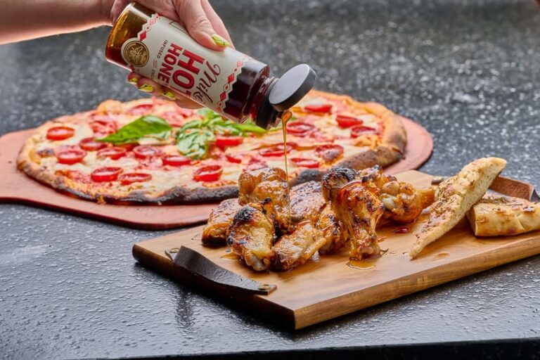 A hand pouring honey over a plate of grilled chicken wings, with a fresh coal-fired pizza in the background.