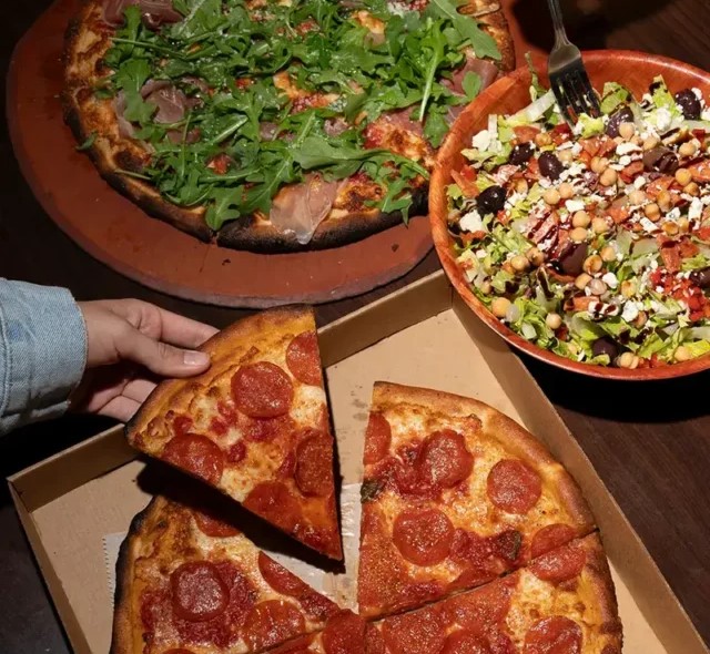 Flat-lay shot of a person taking a slice from Anthony’s Pepperoni Pizza placed beside other dishes.