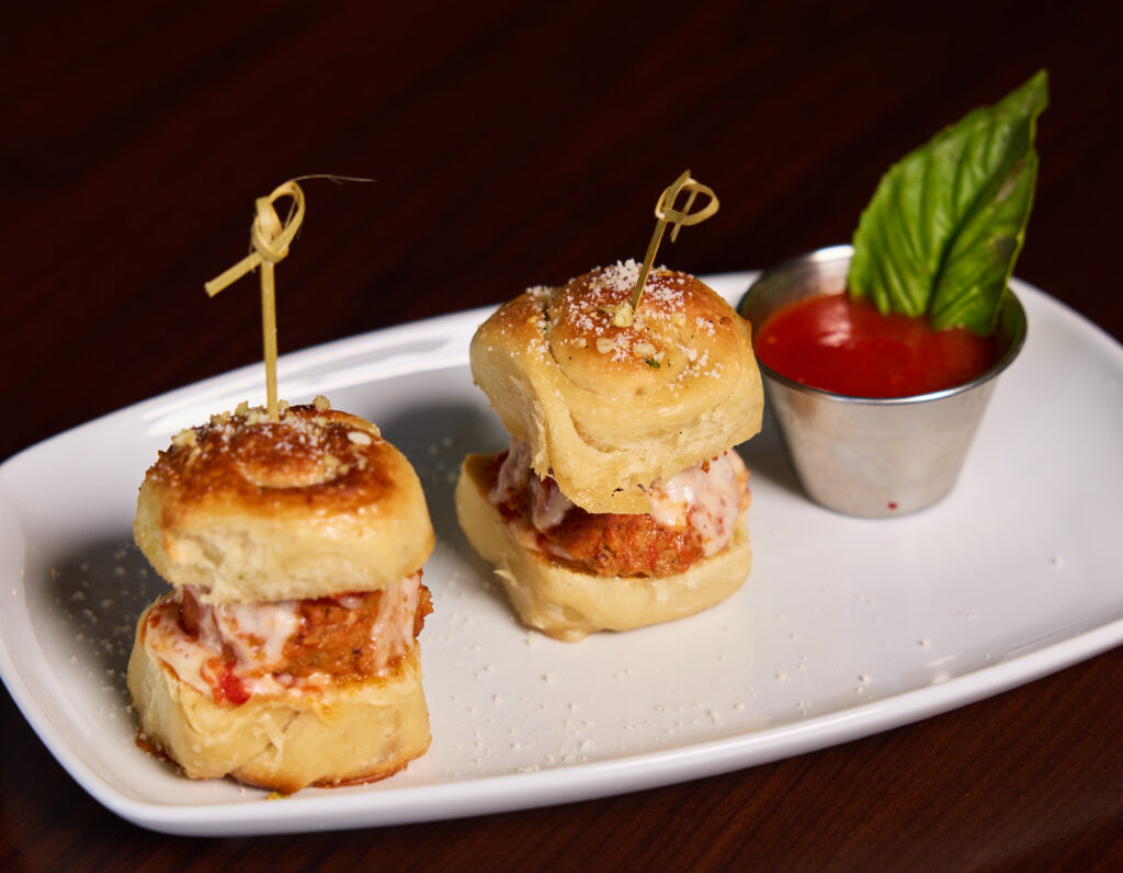 Two mini meatball sliders on spiral roll buns, topped with melted cheese and grated parmesan, served on a white plate with a side of marinara sauce and a basil leaf.