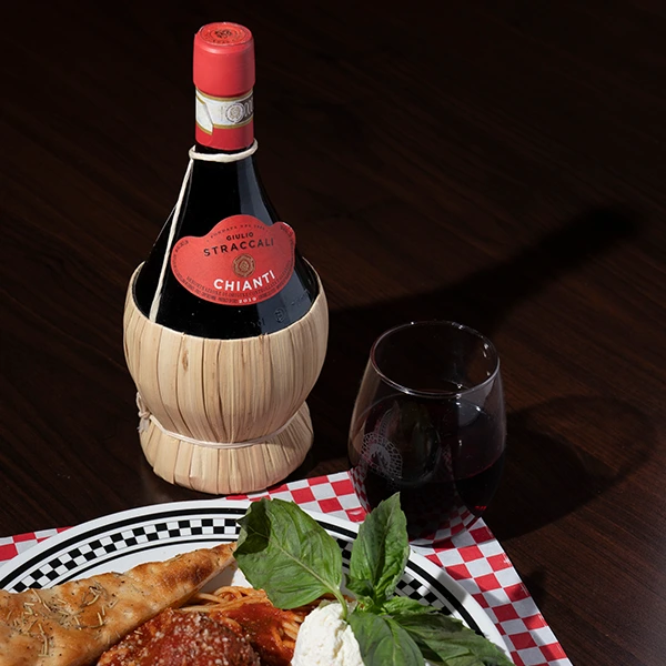 A bottle of red wine and a glass of wine sit beside an Italian meal on a restaurant table.