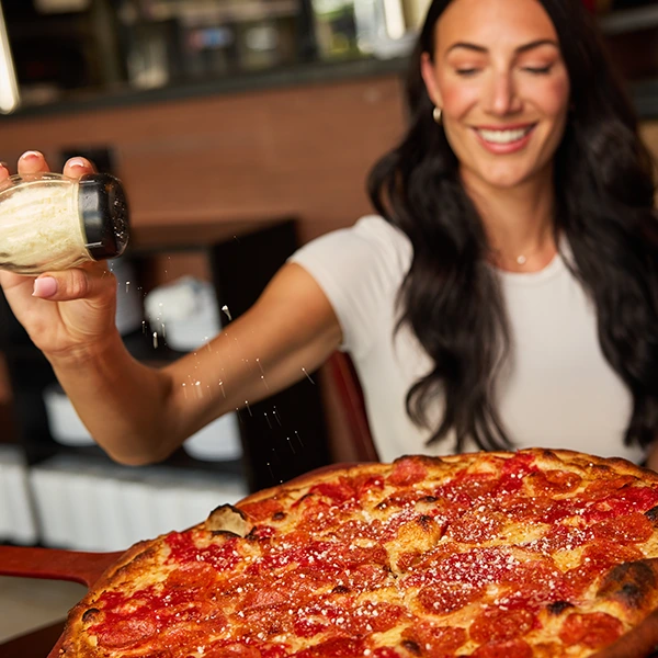 A woman sprinkles grated cheese over a large pepperoni pizza in a restaurant.