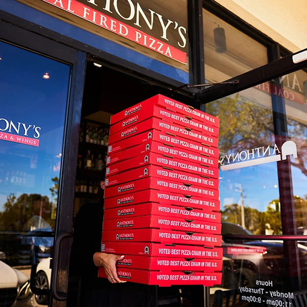 A team member carries a stack of pizza boxes out of an Anthony’s restaurant, highlighting takeout and delivery orders.